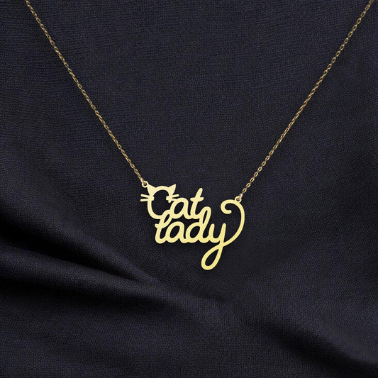 May include: A gold necklace with a pendant featuring the words 'Cat Lady' in a stylized script font. A small cat silhouette is incorporated into the design. The necklace is shown against a dark gray fabric background. The pendant is made of gold metal and has a delicate, feminine design. The chain is a fine, delicate gold chain.