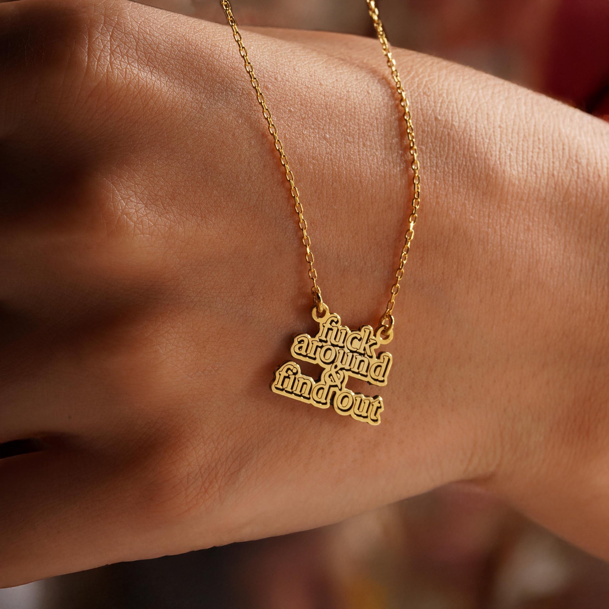 May include: A gold-colored necklace with a pendant that has the words 'fuck around & find out' in a stacked, bold font. The necklace chain is delicate, and the pendant is attached to the chain with small loops. The necklace is displayed against a skin-toned background.
