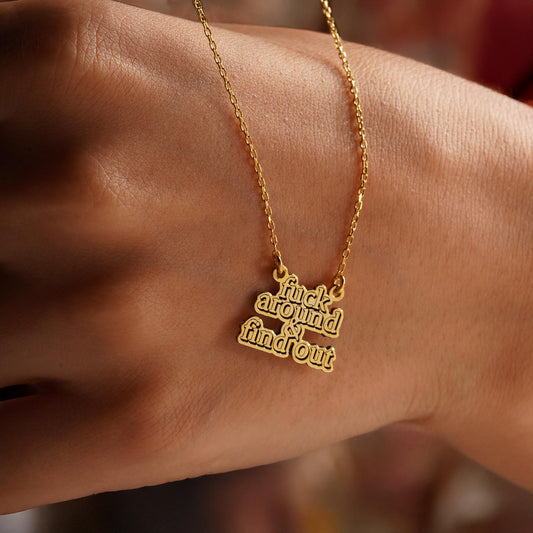 May include: A gold-colored necklace with a pendant that has the words 'fuck around & find out' in a stacked, bold font. The necklace chain is delicate, and the pendant is attached to the chain with small loops. The necklace is displayed against a skin-toned background.