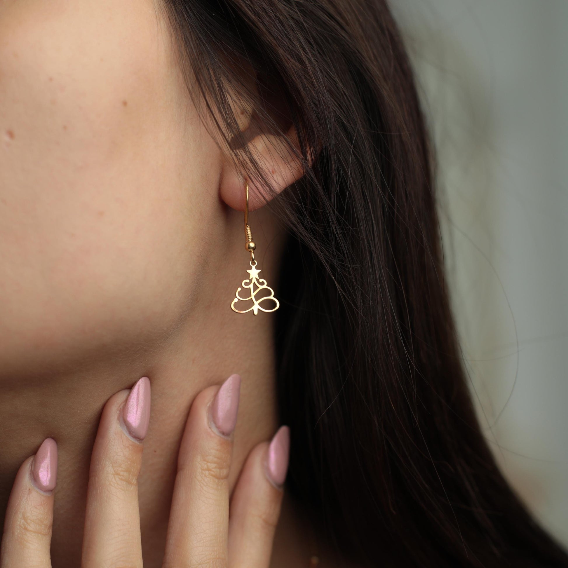 May include: Gold-colored Christmas tree earrings. The earrings are a drop style, with a hook that goes through the ear. The tree design is a cutout, with a star at the top. The earrings are worn by a person with dark hair.