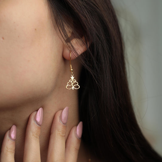 May include: Gold-colored Christmas tree earrings. The earrings are a drop style, with a hook that goes through the ear. The tree design is a cutout, with a star at the top. The earrings are worn by a person with dark hair.