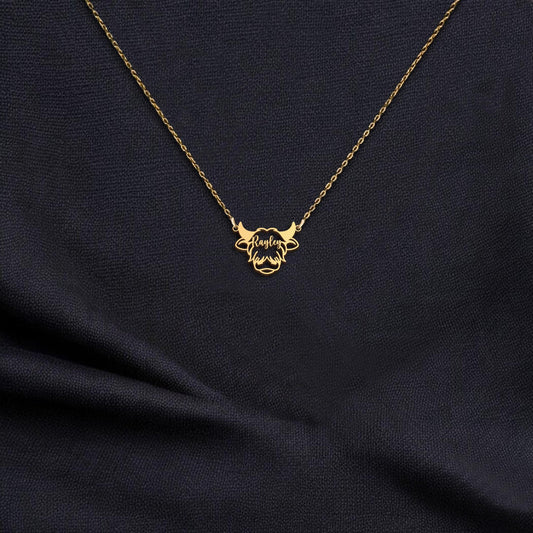 May include: A gold-colored necklace with a pendant shaped like a bull's head. The bull's head has horns and a detailed design. The name 'Raylen' is written in cursive across the center of the bull's head. The necklace is displayed on a dark blue fabric.