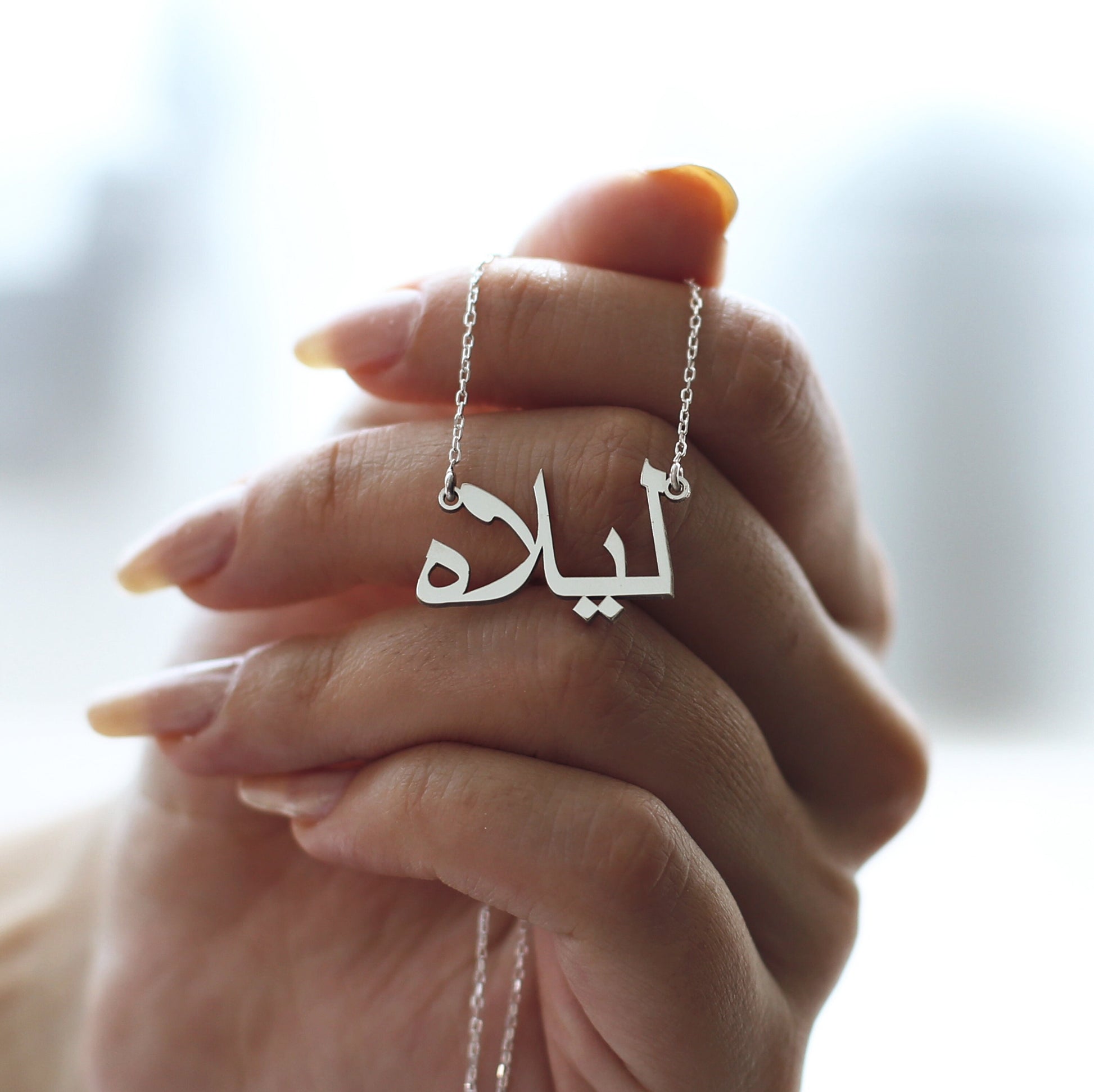 May include: A silver necklace with a pendant featuring the Arabic word "Lila" in a stylized font.